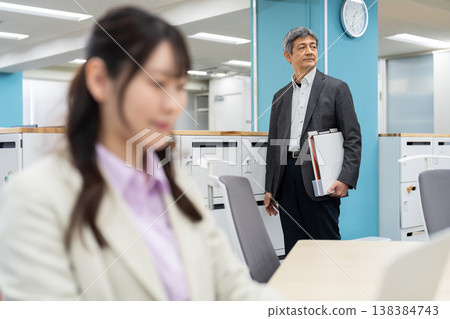A woman is working on her laptop, with a man in the background looking for a seat in a free-address office. A woman is working on her laptop, with a man in the background looking for a seat in a free-address office. 138384743