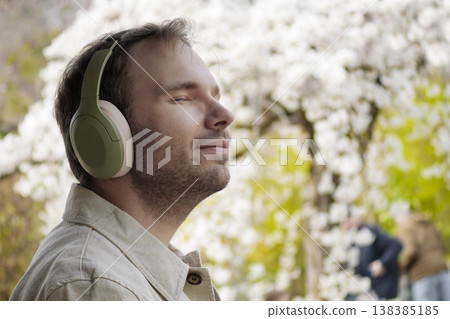 A man wearing headphones relaxes in nature with his eyes closed among blossoming spring trees in a Japanese park. Slow living A man wearing headphones relaxes in nature with his eyes closed among blossoming spring trees in a Japanese park. Slow living 138385185