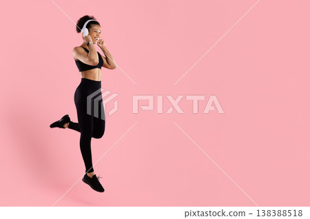 Energetic black woman jumping with headphones in sportswear on pink studio background Energetic black woman jumping with headphones in sportswear on pink studio background 138388518