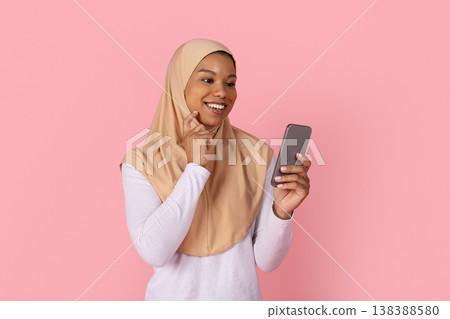 Young black Muslim woman in hijab smiling playfully while holding smartphone Young black Muslim woman in hijab smiling playfully while holding smartphone 138388580