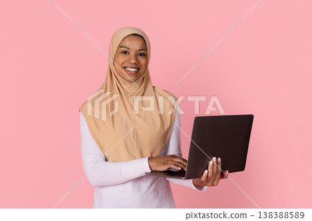 Young black Muslim woman in hijab smiling confidently while holding open laptop 138388589