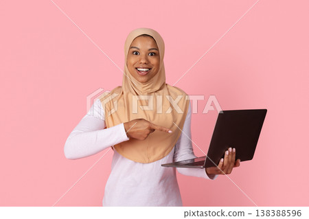 Young black Muslim woman in hijab pointing excitedly at laptop in hands Young black Muslim woman in hijab pointing excitedly at laptop in hands 138388596