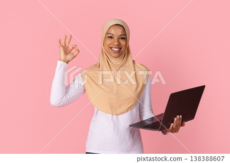 Young black Muslim woman in hijab showing OK gesture while holding laptop Young black Muslim woman in hijab showing OK gesture while holding laptop 138388607