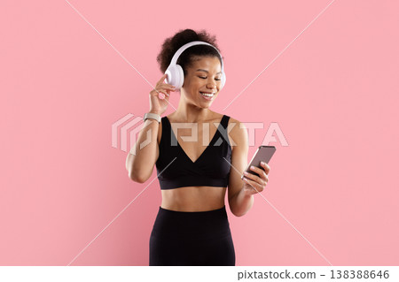 Smiling black sportswoman in activewear adjusting white headphones while looking at smartphone Smiling black sportswoman in activewear adjusting white headphones while looking at smartphone 138388646