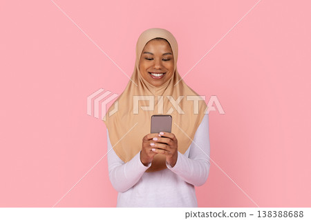 Young black Muslim woman in beige hijab smiling while looking at smartphone Young black Muslim woman in beige hijab smiling while looking at smartphone 138388688