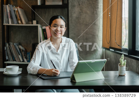 A young woman in a white shirt works on a digital tablet at a desk in a modern office setting with natural light. A young woman in a white shirt works on a digital tablet at a desk in a modern office setting with natural light. 138389729