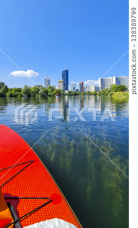 First-person view from a red stand-up paddleboard on the modern Vienna skyline reflected in Danube river. Summer water sport. 138389799