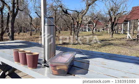 A stainless steel thermos with paper cups and a lunch box on a wooden table in a sunny spring garden A stainless steel thermos with paper cups and a lunch box on a wooden table in a sunny spring garden 138391080