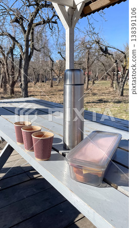 Stainless steel thermos with paper cups and a lunch box on a wooden table in a sunny spring orchard Stainless steel thermos with paper cups and a lunch box on a wooden table in a sunny spring orchard 138391106