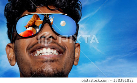 Smiling man in sunglasses with beach volleyball reflected in lenses under blue sky. Smiling man in sunglasses with beach volleyball reflected in lenses under blue sky. 138391852