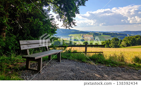 Countryside vacation, summer. wooden bench overlooks scenic countryside landscape with rolling hills and bright blue sky 138392434