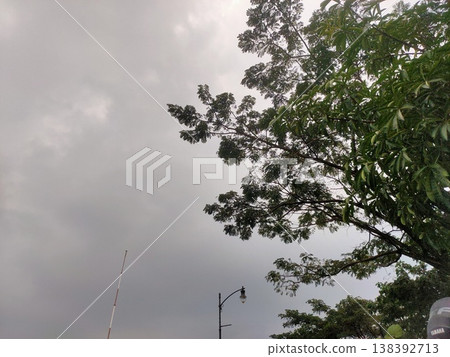 Dark clouds gather above lush trees, hinting at rain on a quiet, overcast afternoon. 138392713