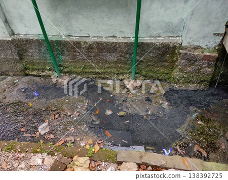 Polluted drainage with dirty water, plastic waste, and moss-covered walls in an urban alley setting. Polluted drainage with dirty water, plastic waste, and moss-covered walls in an urban alley setting. 138392725