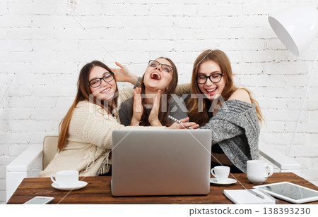Three friends sit together at a wooden table, laughing and engaging with a laptop. They have drinks nearby and are in a light-colored room with white walls. Three friends sit together at a wooden table, laughing and engaging with a laptop. They have drinks nearby and are in a light-colored room with white walls. 138393230