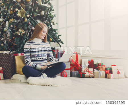 Young woman shopping online on laptop in cozy christmas interior. Girl in casual sitting under decorated pine tree among lots of wrapped presents. Preparing to xmas, bying on winter sales. Copy space 138393395