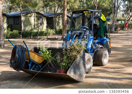 Compact utility loader with gardening tools plants and wheelbarrow in campsite environment maintenance work concept Compact utility loader with gardening tools plants and wheelbarrow in campsite environment maintenance work concept 138393456