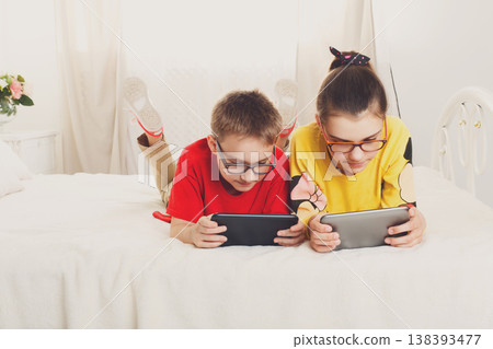 Two children, siblings on parents bed at morning with laptop and tablet. Brother and sister play computer games. Siblings and gadgets. Children with pet, chiwawa dog in bed. Children in glasses. 138393477