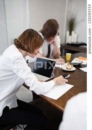 Woman reviewing notes at long wooden office table during company breakfast meeting with papers and coffee. Reviewing, notes, table, breakfast, coffee, office, woman. Woman reviewing notes at long wooden office table during company breakfast meeting with papers and coffee. Reviewing, notes, table, breakfast, coffee, office, woman. 138393554