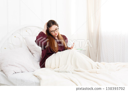 Young waking up woman with tablet pc in white bed. Girl in eyeglasses looks into gadget tablet pc. Morning coffee. Beautiful woman in nightwear at bed surfing internet at tablet computer. High key. 138393702