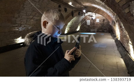 Boy photographing museum exhibits in an underground vault, exploring history with a smartphone. Low light travel scene, child curiosity, education and digital exploration. Boy photographing museum exhibits in an underground vault, exploring history with a smartphone. Low light travel scene, child curiosity, education and digital exploration. 138393719
