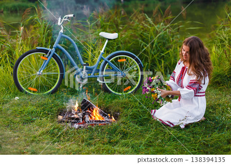 Woman in Folk costume with bicycle. Young attractive woman at riverside having rest with bicycle in national ethnic ukrainian folk traditional dress. Girl looking at circlet of flowers near bonfire. Woman in Folk costume with bicycle. Young attractive woman at riverside having rest with bicycle in national ethnic ukrainian folk traditional dress. Girl looking at circlet of flowers near bonfire. 138394135