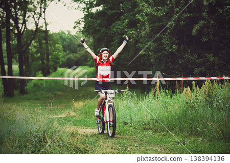 Winner. Victory moment. Young red-haired woman cyclist in protective helmet crossing the finish line breaking the tape. Victory triumph in sportive competition, wins the cycling race. Bicycle sport. Winner. Victory moment. Young red-haired woman cyclist in protective helmet crossing the finish line breaking the tape. Victory triumph in sportive competition, wins the cycling race. Bicycle sport. 138394136