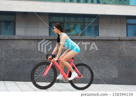 Sexy girl with bicycle. Young slim sexy sporty woman in blue shorts and white snickers long-haired, sensual riding at pink fix bicycle in urban city environment. Soft toned. 138394139