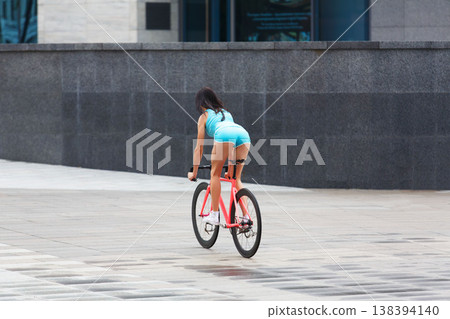 Sexy girl with bicycle. Young slim sexy sporty woman in blue shorts and white snickers long-haired, sensual posing with pink fix bicycle in urban city environment. Soft toned. Shaped buttocks. 138394140