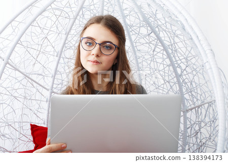 A young woman sits in a white hanging chair holding a laptop. She looks focused and is working on her computer at home in a bright and simple room. A young woman sits in a white hanging chair holding a laptop. She looks focused and is working on her computer at home in a bright and simple room. 138394173