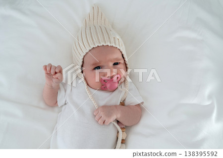 Smiling newborn baby with pacifier lying on soft white bedding. Cozy minimal home setting, concept of infancy comfort and peaceful family life. 138395592