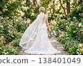 A bride's back view, with her veil fluttering in the wind, in a garden overflowing with blooming roses. 138401049