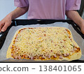 Process of making homemade pizza on a baking sheet with person hands holding tray unbaked dough with tomato sauce ham bell peppers and lots of shredded cheese top view kitchen domestic cooking hobby 138401065