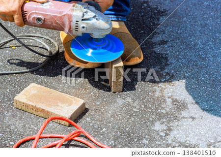 Worker cuts block with grinder tool while standing on dark pavement with other tools, materials around. 138401510