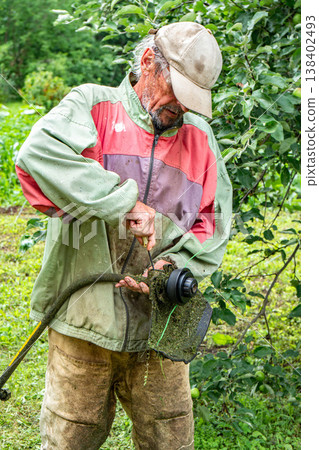 Elderly man adjusts electric grass trimmer while wearing dirty clothes in garden setting 138402493