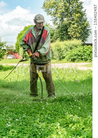 Elderly man in cap cuts grass with electric trimmer in front of his home in the afternoon Elderly man in cap cuts grass with electric trimmer in front of his home in the afternoon 138402497