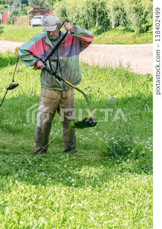 Elderly man mows grass with an electric trimmer in front of his house in residential area Elderly man mows grass with an electric trimmer in front of his house in residential area 138402499
