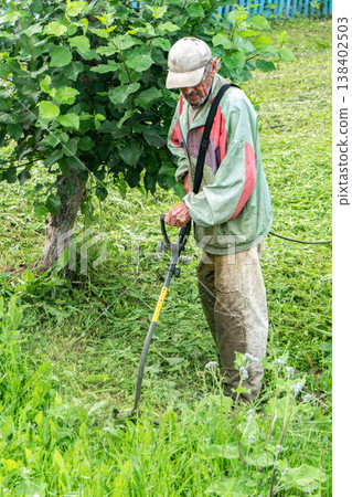 Elderly man uses electric trimmer to cut grass in yard on a sunny day Elderly man uses electric trimmer to cut grass in yard on a sunny day 138402503