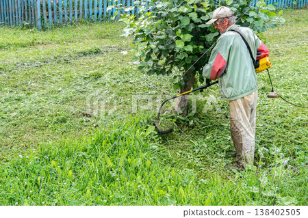 Elderly man mows grass with electric trimmer in front of his home Elderly man mows grass with electric trimmer in front of his home 138402505