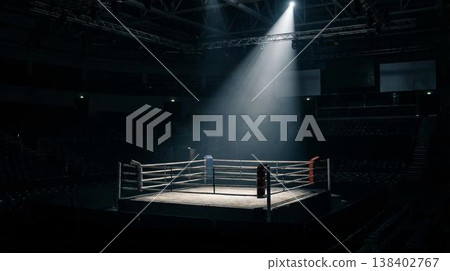 Boxing ring illuminated by a spotlight in an empty arena, with seating visible in the background and a dark atmosphere enhancing the scene's intensity 138402767