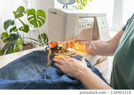 Hands operating a sewing machine, stitching blue denim fabric of jeans, repairing clothing. Person sewing denim jeans on a sewing machine Hands operating a sewing machine, stitching blue denim fabric of jeans, repairing clothing. Person sewing denim jeans on a sewing machine 138403218