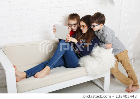 Three children sit on a sofa looking at a tablet together. They seem engaged in a video or games. The setting is bright and inviting with a modern design. Three children sit on a sofa looking at a tablet together. They seem engaged in a video or games. The setting is bright and inviting with a modern design. 138403448