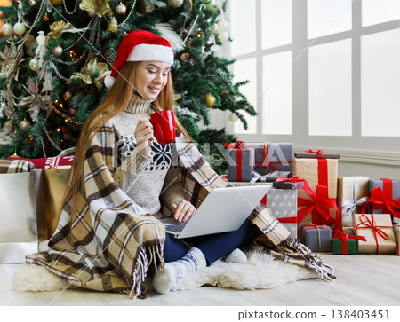 Young woman with coffee shopping online on laptop in christmas interior. Girl sitting under decorated fir tree among lots of wrapped presents. Preparing to xmas, bying on winter sales, copy space Young woman with coffee shopping online on laptop in christmas interior. Girl sitting under decorated fir tree among lots of wrapped presents. Preparing to xmas, bying on winter sales, copy space 138403451