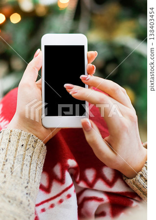 Christmas online shopping. Female buyer makes order at screen of smartphone with copy space. Woman buys presents for xmas eve, sits at home near pine tree with shining lights. Winter holidays sales Christmas online shopping. Female buyer makes order at screen of smartphone with copy space. Woman buys presents for xmas eve, sits at home near pine tree with shining lights. Winter holidays sales 138403454
