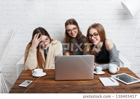 Three friends are sitting around a wooden table in a well-lit indoor area. They are looking at a laptop screen and sharing a moment while enjoying coffee. Three friends are sitting around a wooden table in a well-lit indoor area. They are looking at a laptop screen and sharing a moment while enjoying coffee. 138403457