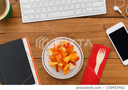 Healthy lunch at workplace. Top view flat lay of desktop fruit snack in foil container and mobile phone on wooden desk background Healthy lunch at workplace. Top view flat lay of desktop fruit snack in foil container and mobile phone on wooden desk background 138403462