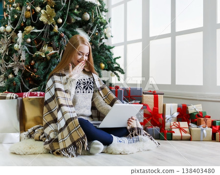 Happy girl with laptop sitting at decorated christmas tree background. Woman got greeting email from online shop with huge discount at winter sale. Holiday atmosphere, copy space Happy girl with laptop sitting at decorated christmas tree background. Woman got greeting email from online shop with huge discount at winter sale. Holiday atmosphere, copy space 138403480