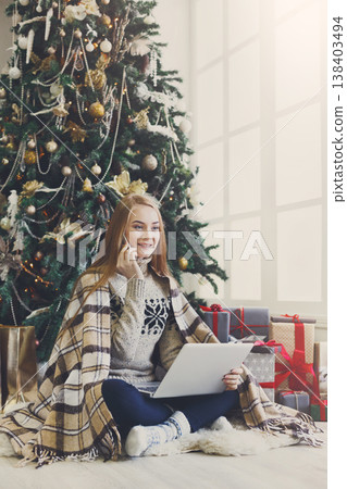 Young woman shopping online on laptop, making order on mobile. Girl sitting under decorated christmas tree among lots of wrapped presents. Preparing to xmas, bying on winter sales. Copy space Young woman shopping online on laptop, making order on mobile. Girl sitting under decorated christmas tree among lots of wrapped presents. Preparing to xmas, bying on winter sales. Copy space 138403494