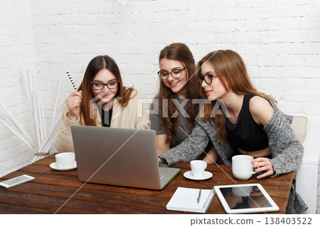 Three friends sit at a wooden table with cups of coffee and devices. They focus on the laptop screen, sharing ideas and working together. The room has a simple design with white walls. 138403522