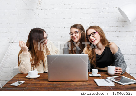 Three friends sit at a wooden table in a bright space. They share smiles and coffee while looking at a laptop. Their casual attire shows a relaxed atmosphere during their meeting. Three friends sit at a wooden table in a bright space. They share smiles and coffee while looking at a laptop. Their casual attire shows a relaxed atmosphere during their meeting. 138403523