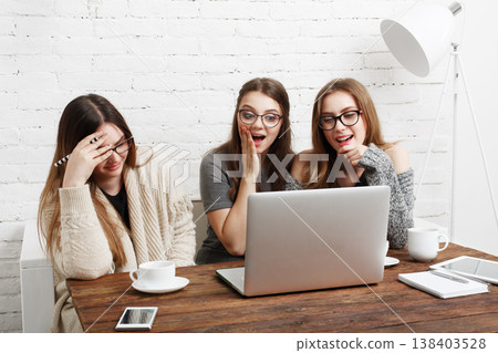 Three friends sit together at a wooden table, reacting with surprise as they watch something on a laptop. They have cups of coffee and a phone on the table in a bright room. 138403528